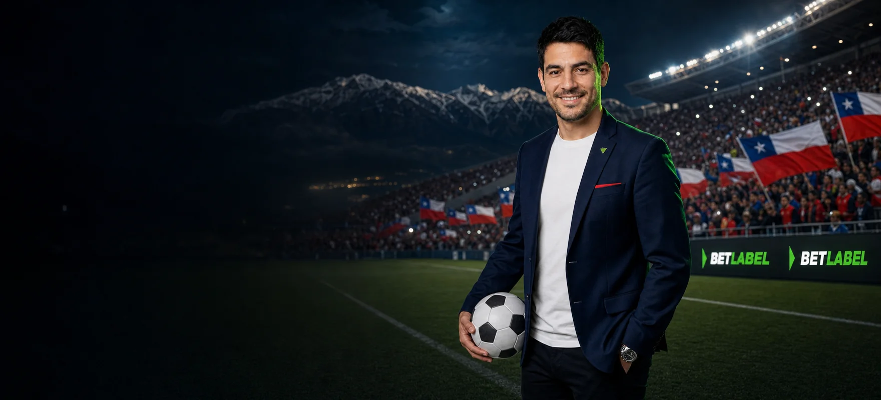 Hombre de chaqueta azul con una pelota junto a la cancha de un estadio iluminado de Santiago, con la cordillera de los Andes al fondo y el logo de BetLabel en las pantallas LED Hombre de chaqueta azul con una pelota junto a la cancha de un estadio iluminado de Santiago, con la cordillera de los Andes al fondo y el logo de BetLabel en las pantallas LED